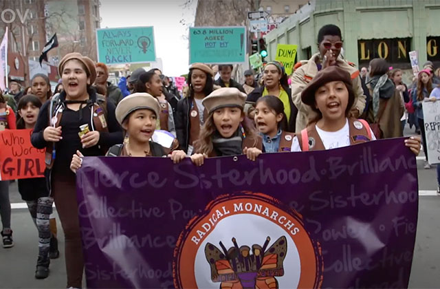 We Are the Radical Monarchs. Three young girls of color wearing scouts caps and carrying a purple banner reading "Radical Monarch" march in a rally.