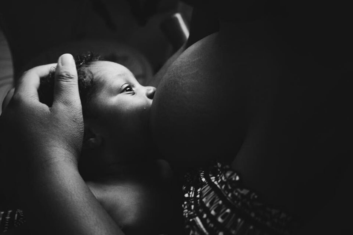 Black and white photo showing a brown baby at a mother's breast, feeding.