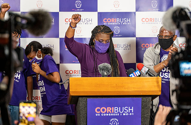 Cori Bush. Black woman wearing purple mask and top with a raised fist a podium surrounded by cameras.