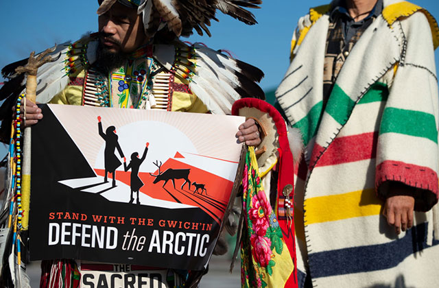 Defend the Arctic Protest. Indigenous man standing next to someone in colorful stripped coat, holds sign reading: Defend the Arctic.