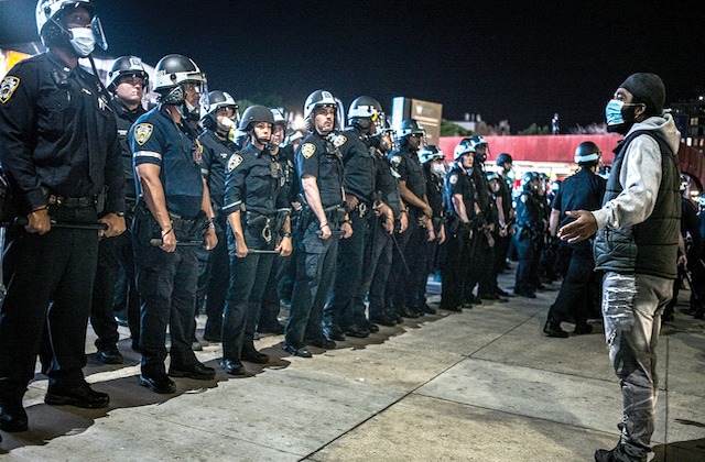 A Black protester wearing a mask stands in front of a blockade of police officers in riot gear.
