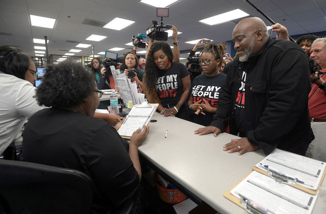 One Black man and two black women stand in front of a desk waiting on paperwork while wearing t-shirts that read, Let My People Vote.