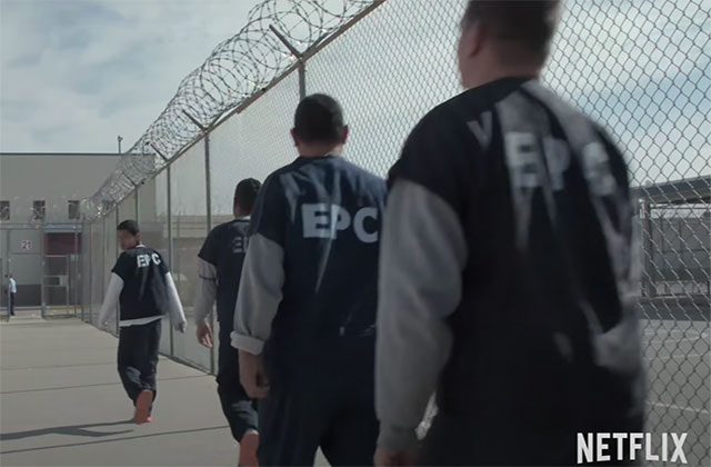 Immigration Nation. Men wearing gray uniforms in single file with "EPC" on the back on shirts and barbed wire overhead.