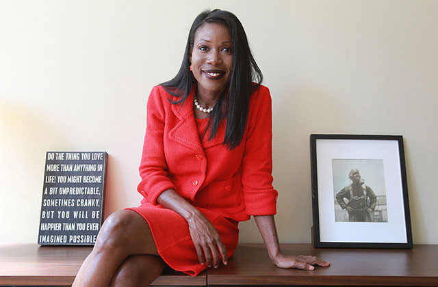 Isabel Wilkerson. Black woman with long dark straight hair wearing red skirt suit sitting between two small pieces of desk art,