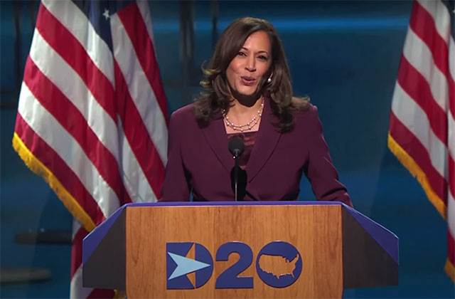 Kamala Harris. Black woman with shoulder length brown hair wearing maroon-colored suit standing in front of U.S. flags on stage.