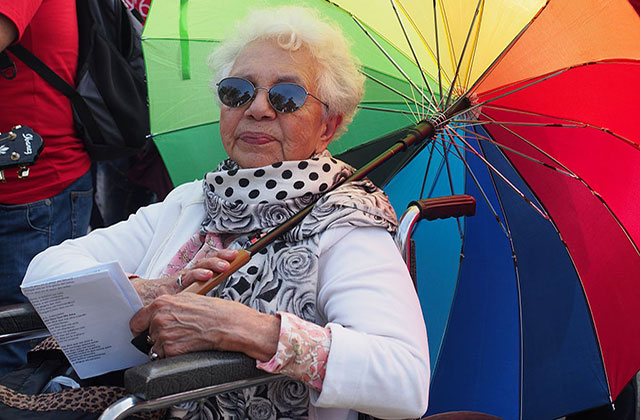 LGBTQ ELDER. Brown elder woman in a wheelchair with short gray hair holding a rainbow colored umbrella.
