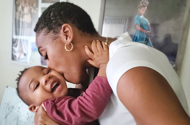 Lyft. Black woman with short dark hair wearing white Tee holding Black baby boy wearing maroon shirt.