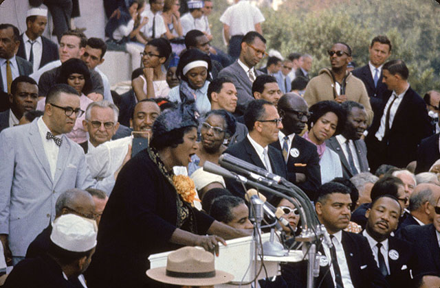 Mahalia Jackson. Black woman on stage in front of microphone surrounded by men in suits, including Martin Luther King, Jr.