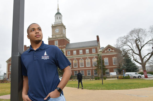 young Black man leans on a poll and looks at the sky as the Howard University campus sits in the background.