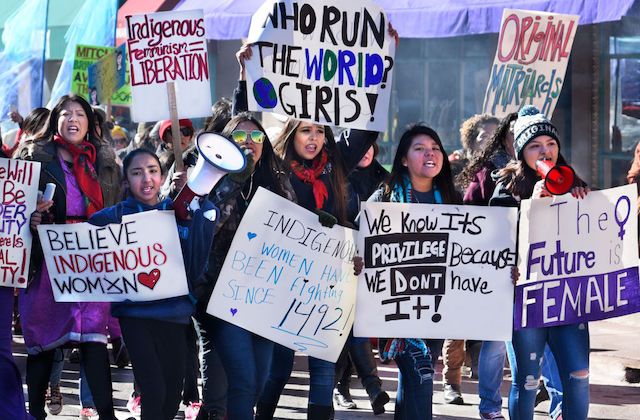 A group of Indigenous women march and hold colorful signs calling for justice. 