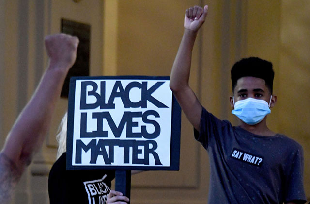 Protest. Young Black boy wearing face mask and blue Tee shirt stands with raised fist in front of sign reading: Black Lives Matter.