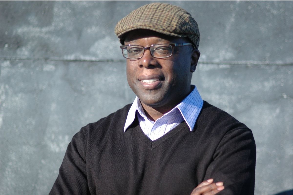 Middle age Black man smiling with paperboy cap, black sweater, and dress shirt underneath