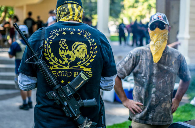 white man with his back to the camera wears a jacket that reads Columbia River Proud Boys. Another white man facing the camera has a yellow bandana covering his mouth and nose.