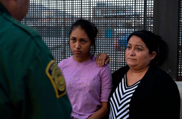 a young girl wearing a purple shirt stands next to her mother. They both look somber as they face a customs and border patrol agent, who stand with his back to the camera. 