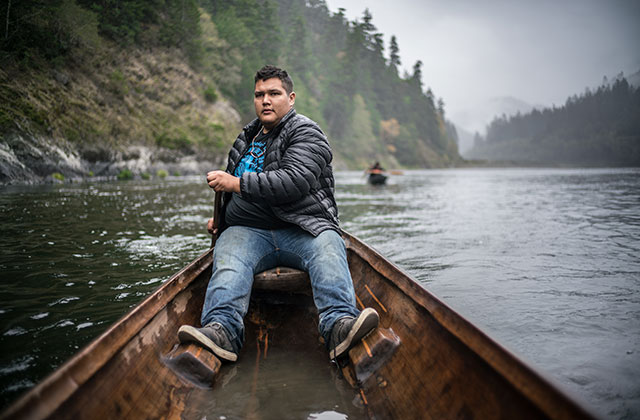 Gather. Indigenous man wearing a black bubble jacket, blue jeans and boots, rowing a boat on a river.
