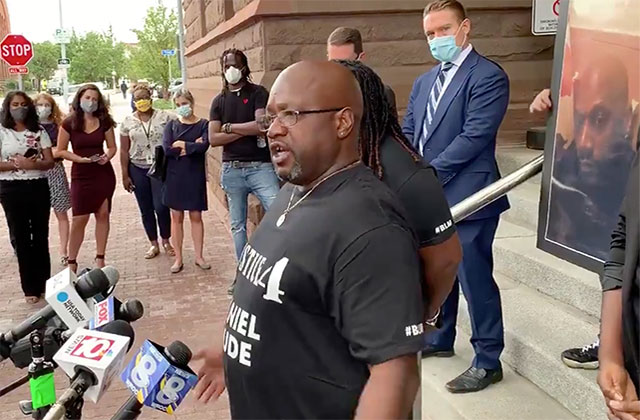 Joseph-Prude. Black man with bald head, glasses wearing black Tee stands in front of reporter's mics.