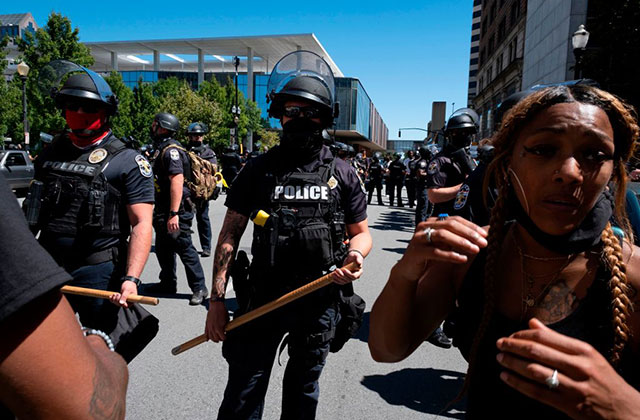 Kentucky-Protests. Black woman with long braids wearing black tank and black face mask moves ahead of a row of police officers holding batons.