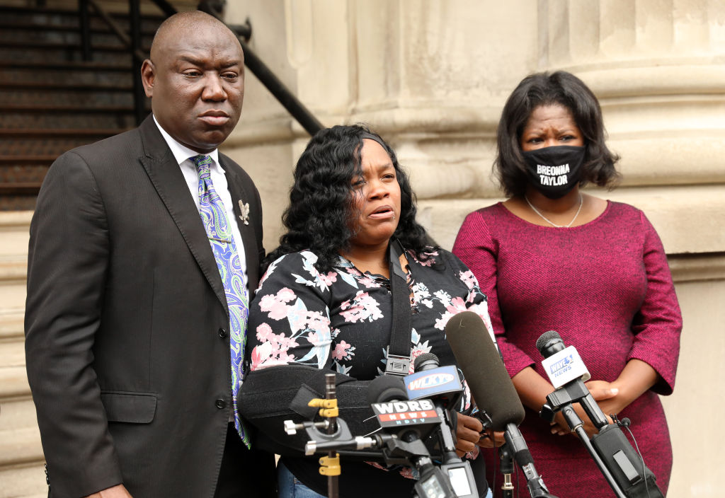 A Black man and two Black women, one wearing a mask that says Breonna Taylor, stand in front of a microphone. 