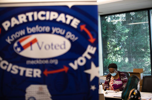 NC-ballot-counter. Black woman in background wearing blue mask and glasses at a desk with a sign in the foreground reading: Participate, Register, Vote.