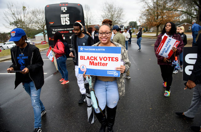 A young Black woman holds a sign that reads Black Votes Matter, as young people march and hold signs behind her.