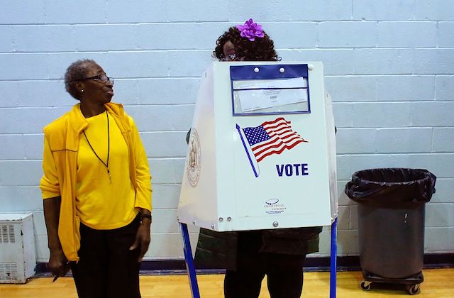 A Black woman wearing a yellow shirt and blazer speaks to another Black woman who wears a purple flower in her hair and stands behind a voting booth.