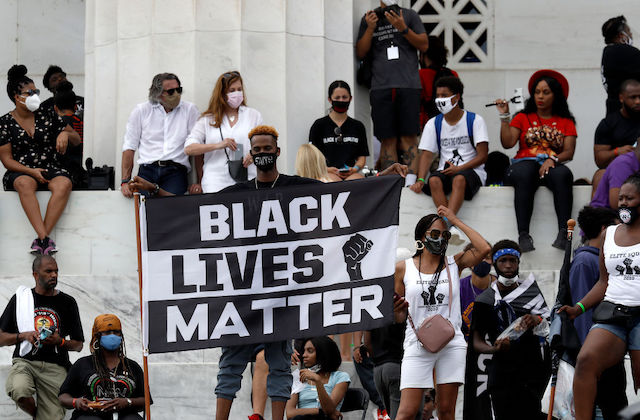 A large group of people are seated on a platform behind a Black man who stands and holds a large Black Lives Matter sign.