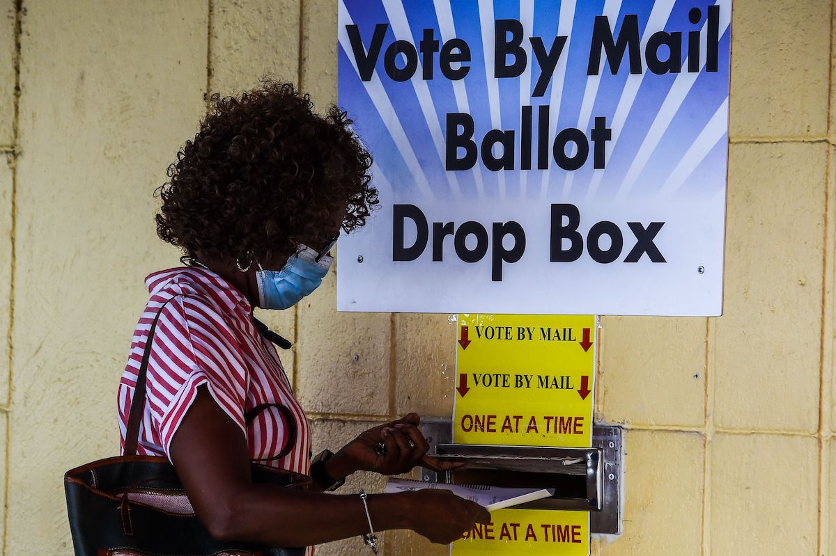A Black woman drops a paper ballot in a slot under colorful yellow and blue signs