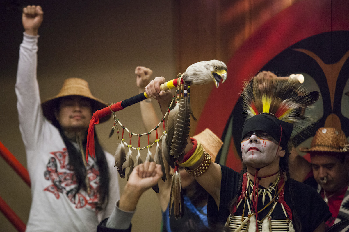 A Native man with painted face and intricate headdress holds aloft a staff and he is surrounded by other Indigenous Americans raising fists in statements of power.