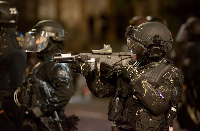 Police officers in black riot gear stand in the middle of a street as one points a gun in front of them.
