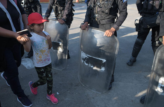 A small girl wearing a red hat hold hands with an adult as they walk past officers wearing riot gear.