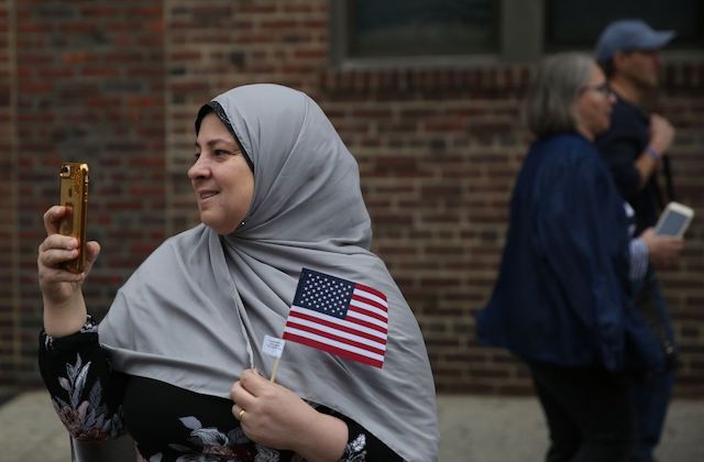 A woman wears a gray hijab smiles as she holds a small American flag and a smartphone.