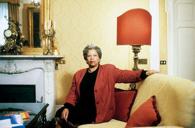 Toni Morrison. Black woman with long gray locs seated on a yellow sofa, wearing a black dress and red jacket.