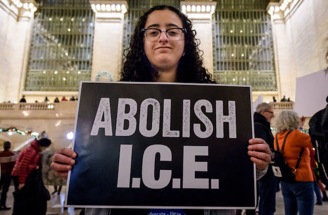 A woman with dark curly hair holds a large sign that says, Abolish ICE