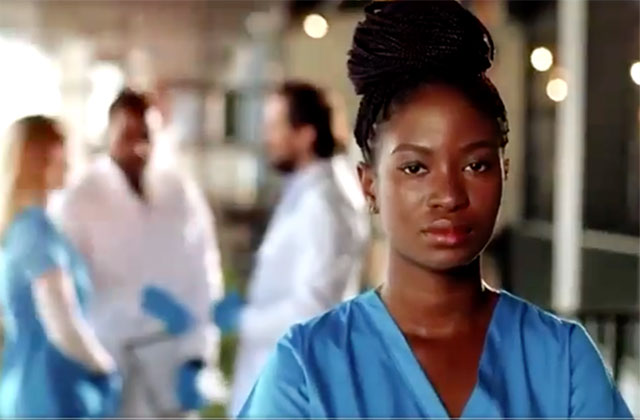 Black Coalition of docs. Young Black woman with braids in top bun wearing blue medical scrubs.