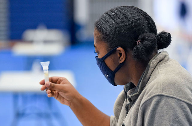 COVID-19 Disparities. Black woman with dark hair in two buns wearing a black mask and gray top holding a vial of saliva.