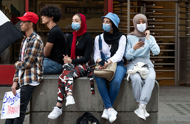 Hijabs. Three women in masks sit on a cinderblock next to two young men without masks.