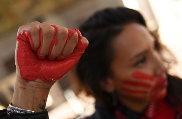 Indigenous Woman. Raised fist painted red with dark-haired woman with mouth covered in red hand in the background.