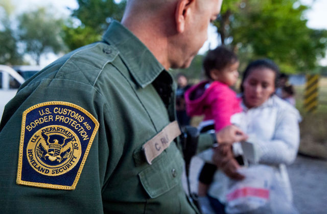 Foreground shows the arm of a man in a green shirt with a patch that reads, US Customs and Border Protection. The background shows a woman holding a small child wearing a pink jacket.