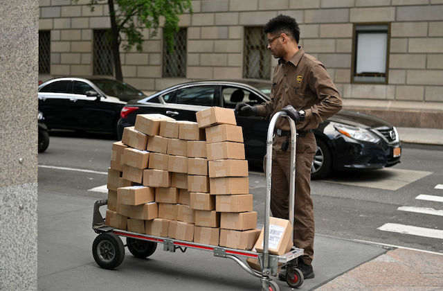 A black man wearing a brown UPS uniform pushes a dally filled with brown, boxed packaged.