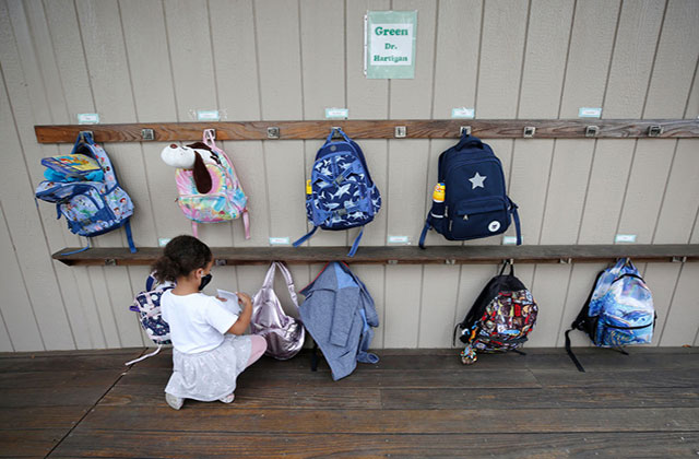LA Schools. Small girl child kneels on floor in front of a row of back packs hanging from a wall.