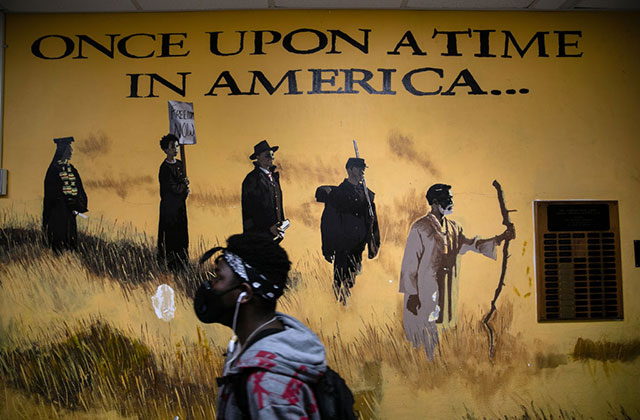 Stamford CT High School. Black male student wearing bandana, black face mask, hoodie and backpack walks by wall mural reading "once upon a time in America."