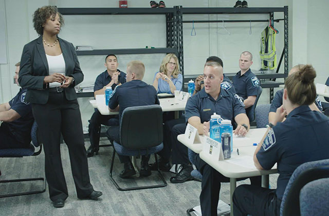 An African American woman in a suit teaches a class of uniformed police officers