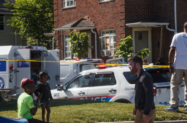 A woman sits with her child behind police tape watching a shooting investigation unfold.