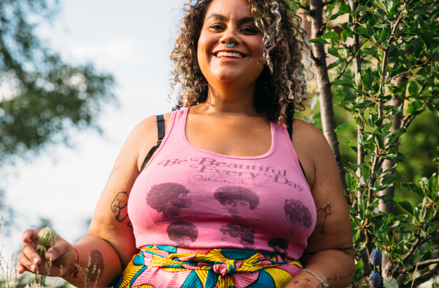 A photo of adrienne maree brown in a pink tank-top that reads "Be Beautiful Everyday"