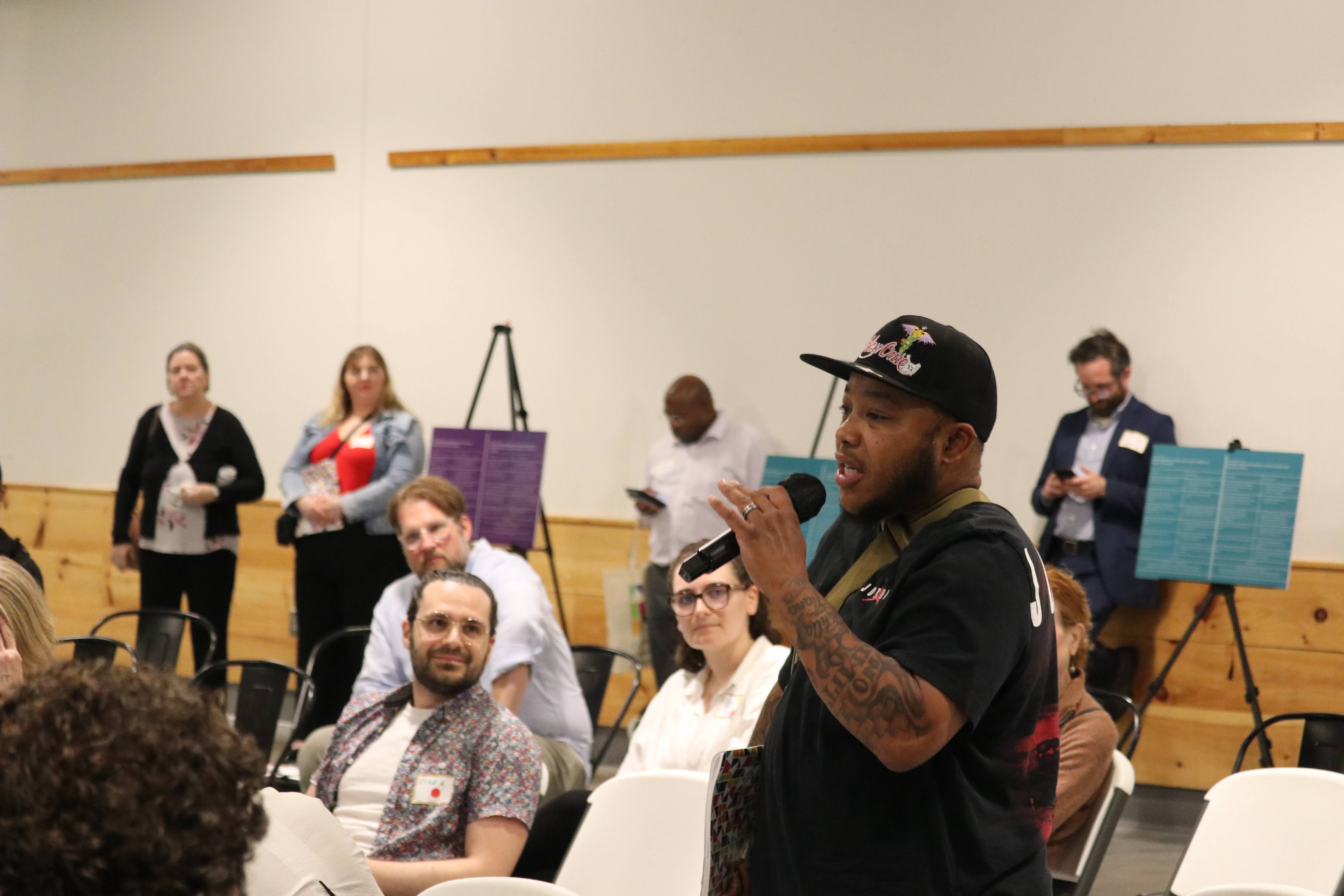 A Black man wearing a black shirt and hat is standing and speaking on a microphone in the middle of a small group while a group of participants look up at him paying close attention.
