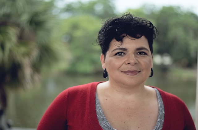 Rosana Cruz. Latinx person in red top in front of green foliage.