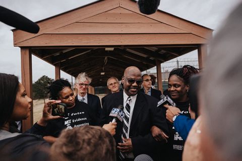 Curtis Flowers. Black man with bald head, dark glasses, dark suit, white shirt and black-and-white tie surrounded by people and reporters.