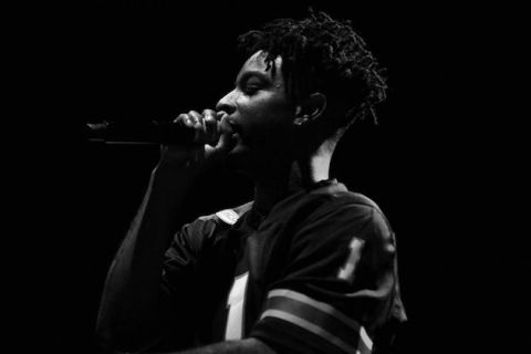21 Savage. Black and white image of Black man holding a microphone and performing on stage. 