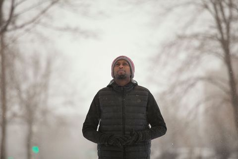 Black man with beard standing in snowy, tree-lined street wearing knit cap, black winter coat, with hands in pockets.