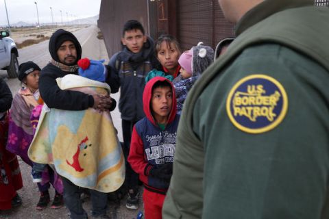 A group of adults and small children gather in front of a a guard wearing a green Customs and Border Protection uniform. 